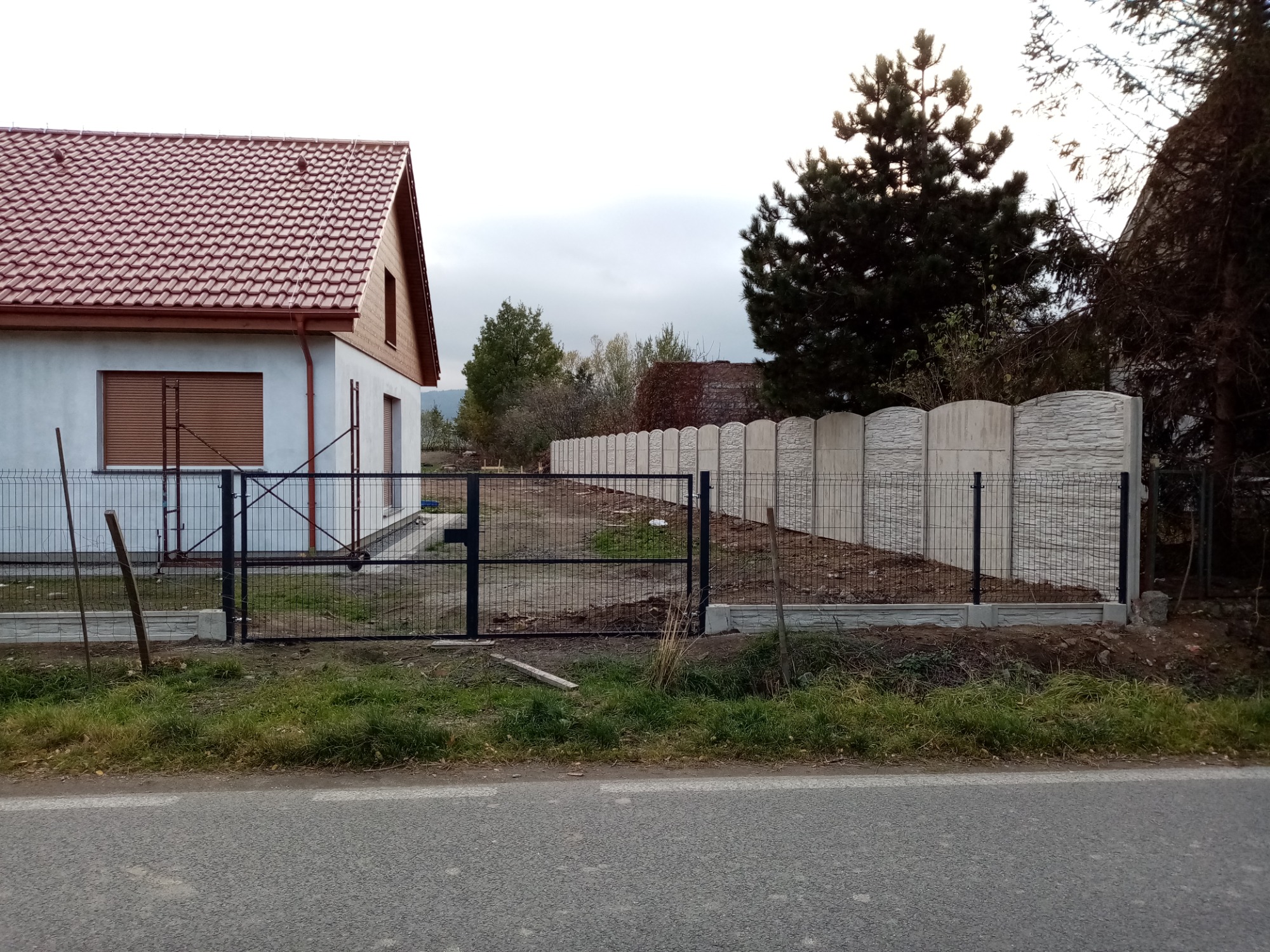 Nowoczesne ogrodzenie z paneli siatkowych i betonowych przęseł, widok z ulicy na posesję z domem w tle, słoneczny dzień.