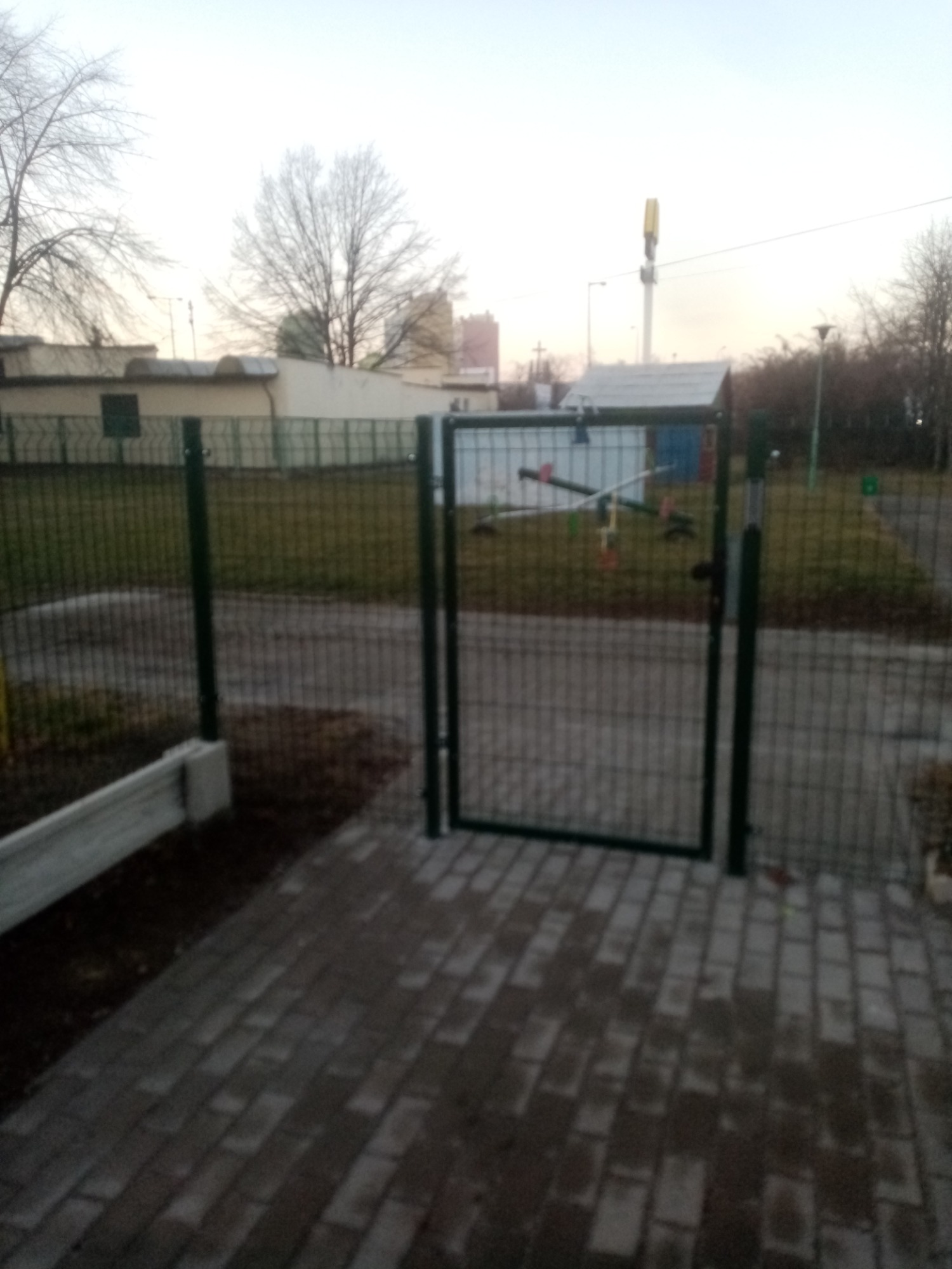 Otwarta furtka z zielonej siatki ogrodzeniowej, prowadząca na plac zabaw z huśtawką, widoczna z perspektywy ułożonej kostki brukowej, w tle budynki i drzewa bez liści.