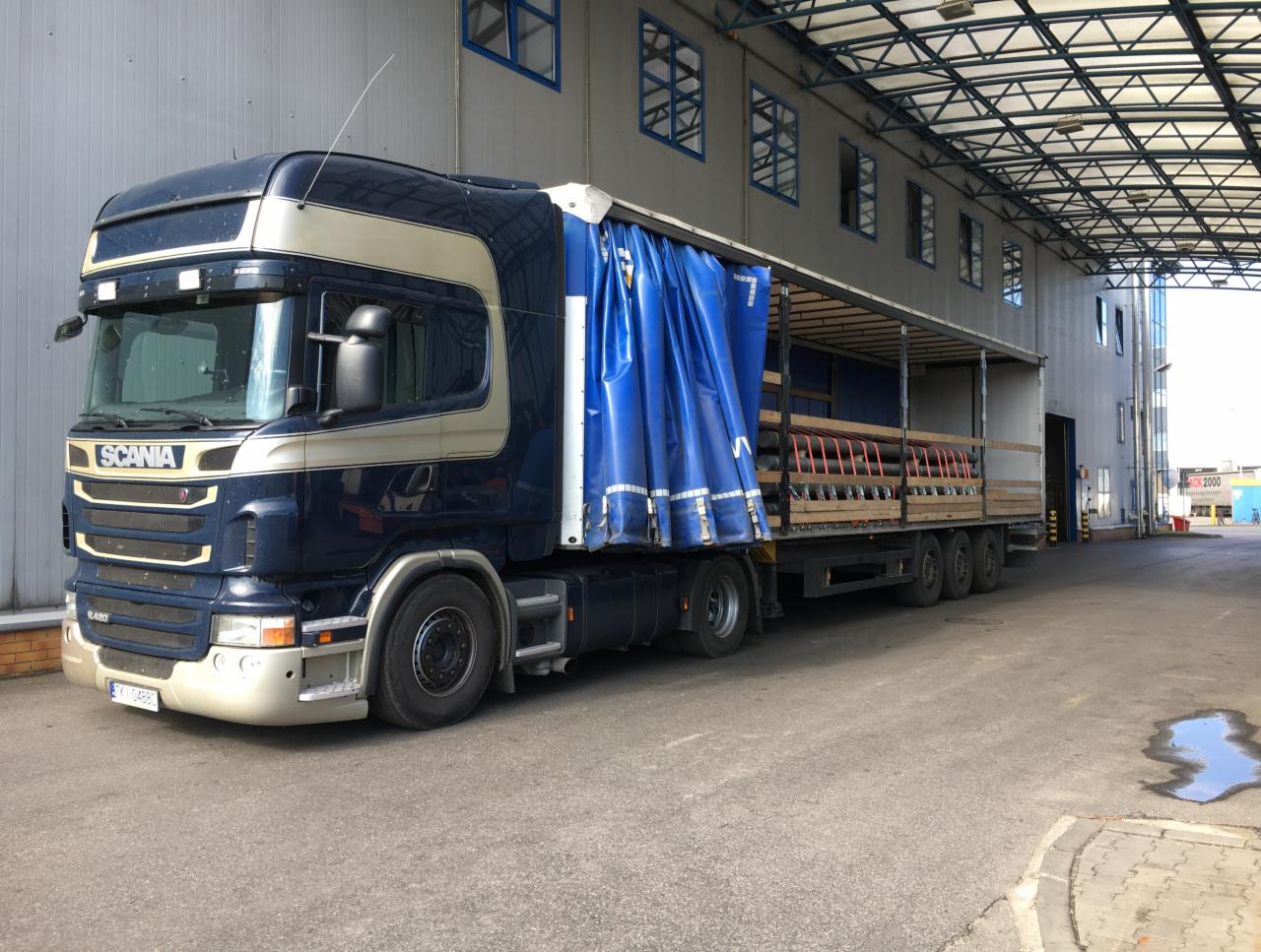 Niebiesko-beżowa ciężarówka Scania R420 z częściowo odsłoniętą plandeką, załadowana rurami, zaparkowana pod zadaszeniem magazynu.