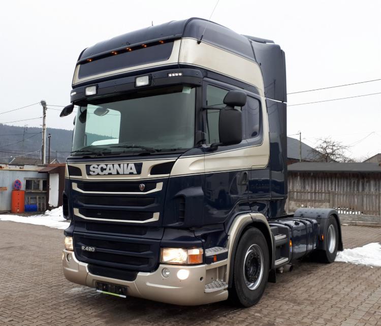 Ciężarówka Scania R420 w kolorze granatowo-beżowym, zaparkowana na brukowanej powierzchni, z widocznymi elementami otoczenia, takimi jak budynek i resztki śniegu.