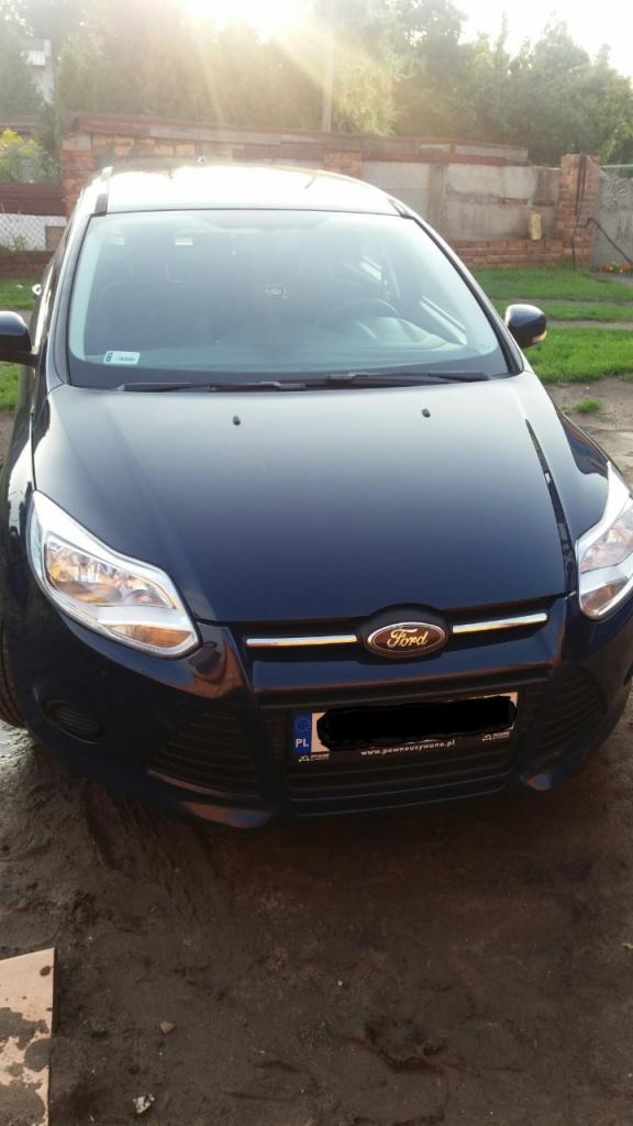 Ciemnoniebieski Ford Focus z widoczną tablicą rejestracyjną i logo marki na tle trawnika i ceglanego muru.