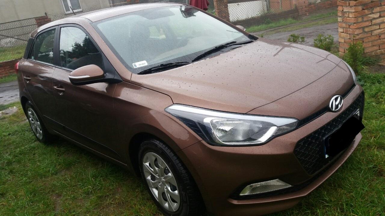 Brązowy samochód osobowy marki Hyundai i20, sfotografowany z zewnątrz, na trawniku przed ceglanym budynkiem w pochmurny dzień.