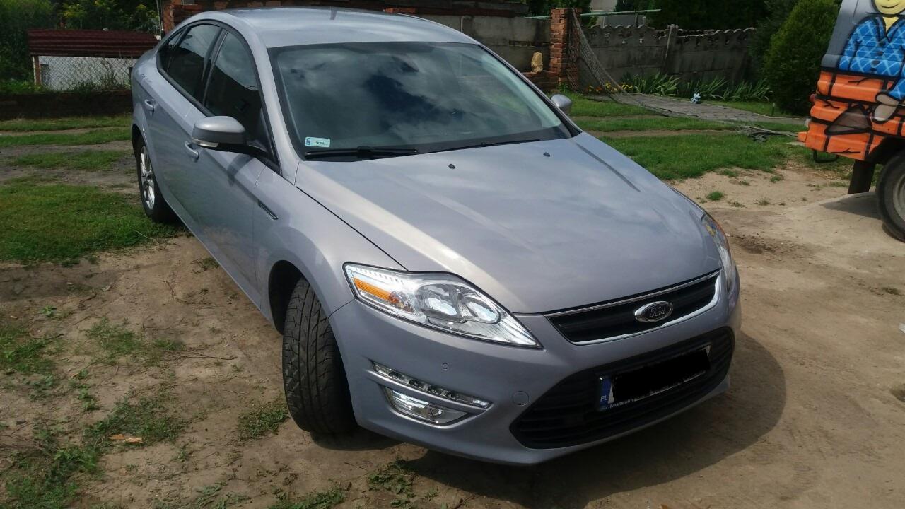 Srebrny Ford Mondeo zaparkowany na ziemnej powierzchni, z widoczną tablicą rejestracyjną i fragmentem kolorowego samochodu w tle.