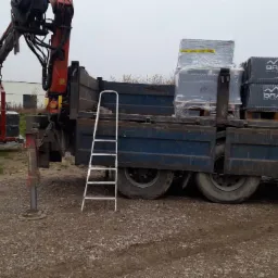 Rozładunek bloczków Braas na budowie domu z użyciem dźwigu samochodowego. Widoczna drabina i fragment surowego muru.