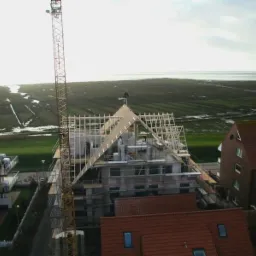 Budowa domu wielorodzinnego z widocznym szkieletem drewnianym dachu, rusztowaniami i żurawiem na tle nadmorskiego krajobrazu.