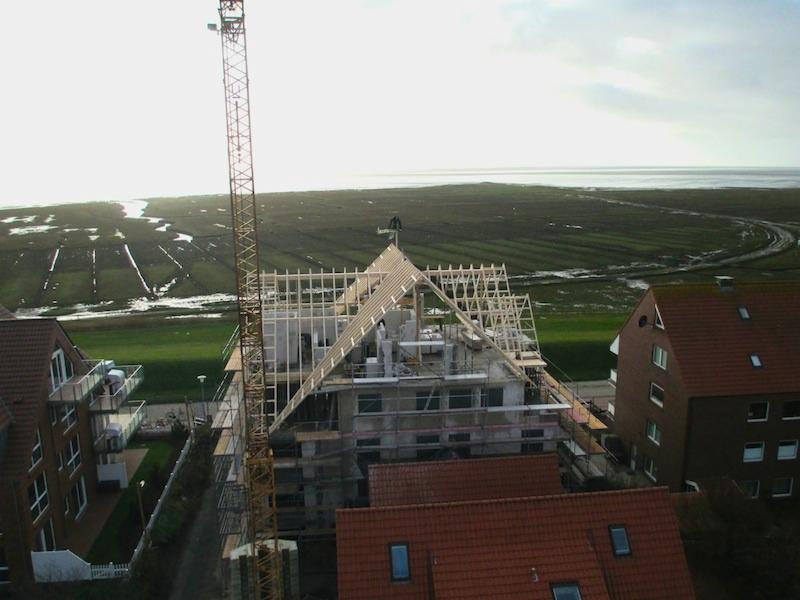 Budowa domu wielorodzinnego z widocznym szkieletem drewnianym dachu, rusztowaniami i żurawiem na tle nadmorskiego krajobrazu.