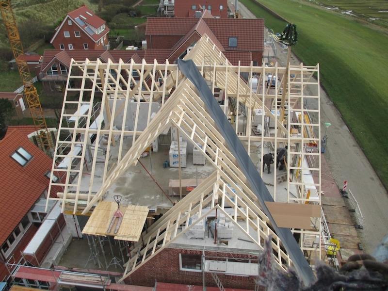 Wysoki kąt widzenia na budowę domu z drewnianą konstrukcją szkieletową dachu, częściowo pokrytą czarną membraną, z widocznym dźwigiem i okoliczną zabudową.