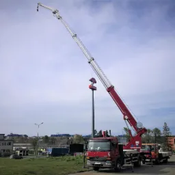 Czerwony dźwig samochodowy z wysuniętym ramieniem ustawia znak stacji paliw Orlen na tle błękitnego nieba z chmurami.