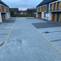 Nowoczesna nawierzchnia z szarej kostki brukowej z żółtymi i czarnymi akcentami, tworząca parking przed nowymi budynkami mieszkalnymi z drewnianymi panelami.