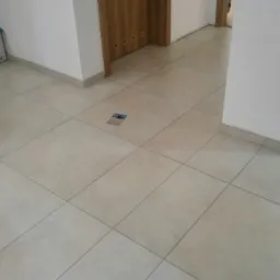 Beżowe płytki podłogowe ułożone w kwadraty, widok na fragment korytarza z białymi ścianami, drewnianymi drzwiami i elementami wyposażenia.