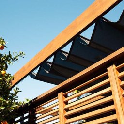 Artbegstudio- - Drewniana konstrukcja z żaluzjami fasadowymi i zwijaną markizą przeciwsłoneczną, widok z dołu na tle błękitnego nieba i drzewa pomarańczowego.