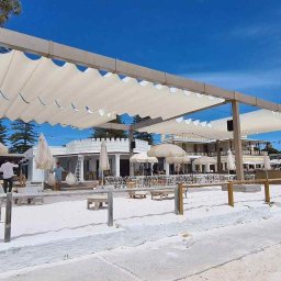 Artbegstudio- - Biała, falista konstrukcja zacieniająca taras restauracji na plaży, widok na błękitne niebo i drzewa w tle.