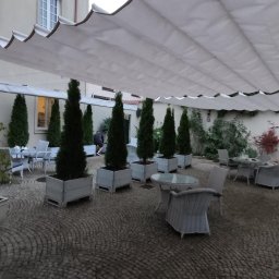 Artbegstudio- - Biała, falista osłona przeciwsłoneczna nad patio z brukowaną nawierzchnią, otoczonym zielenią i wysokimi tujami w kwadratowych donicach.