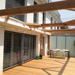 Artbegstudio- - Zewnętrzny taras z drewnianą pergolą i białymi osłonami przeciwsłonecznymi nad oknami zabezpieczonymi metalowymi kratami.