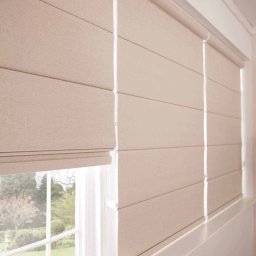 Artbegstudio- - Beżowe rolety rzymskie zamontowane w oknie z widokiem na zielony ogród, częściowo opuszczone.