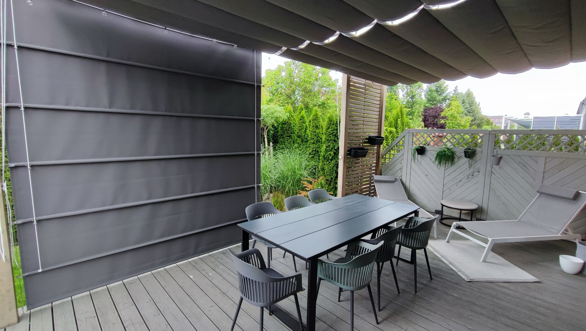 Zacieniony taras z szarą pergolą, zasłonięty z boku ciemnoszarą tkaniną na szynach, stół z krzesłami i leżaki, widok na zielony ogród.