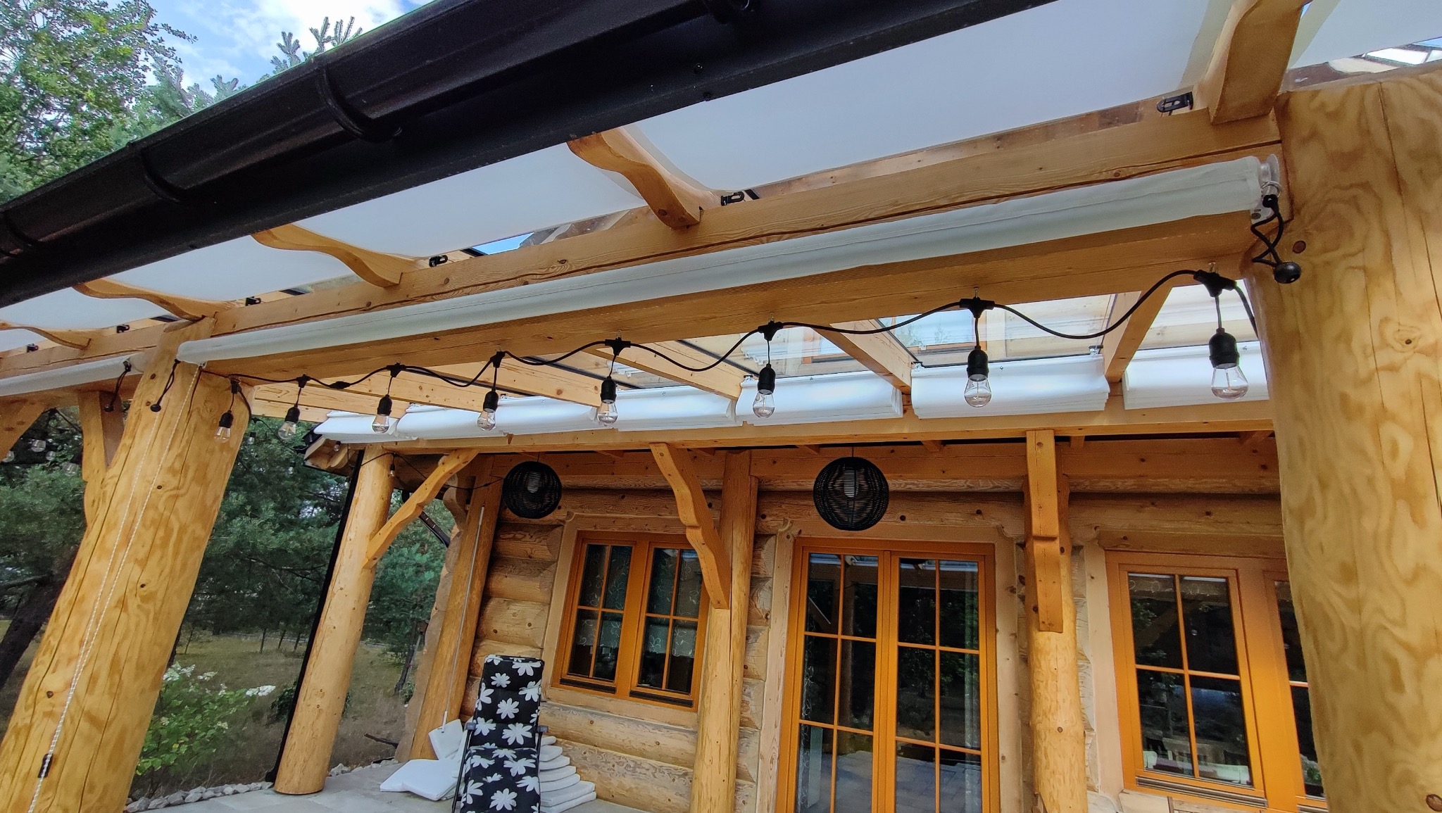 Zadaszenie tarasu z drewnianą konstrukcją, oświetleniem w postaci żarówek i częściowo zwiniętymi materiałowymi osłonami przeciwsłonecznymi.