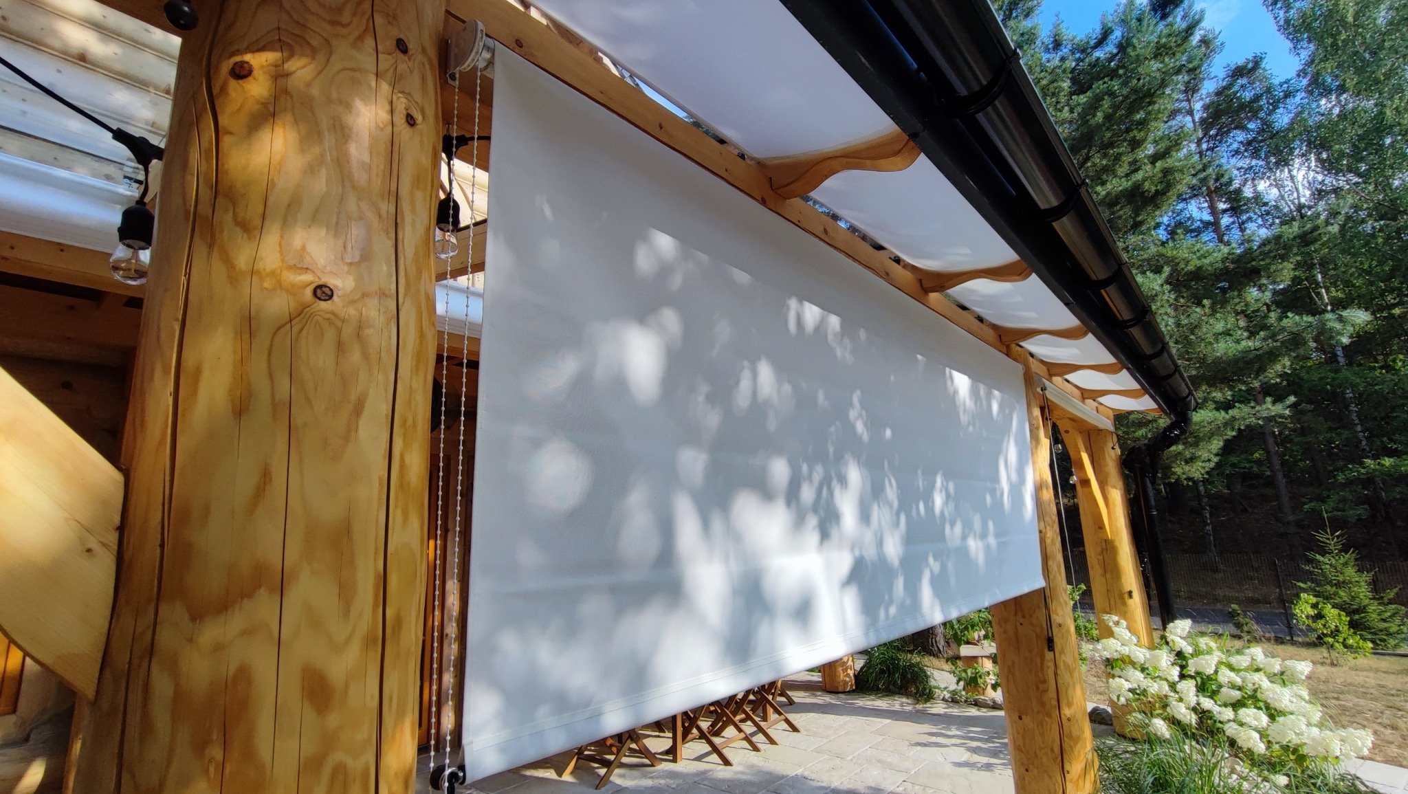 Biała, zwijana przesłona na drewnianej pergoli, z widocznymi elementami mocującymi i łańcuszkiem do regulacji, zacieniająca przestrzeń z krzesłami i roślinnością w tle.