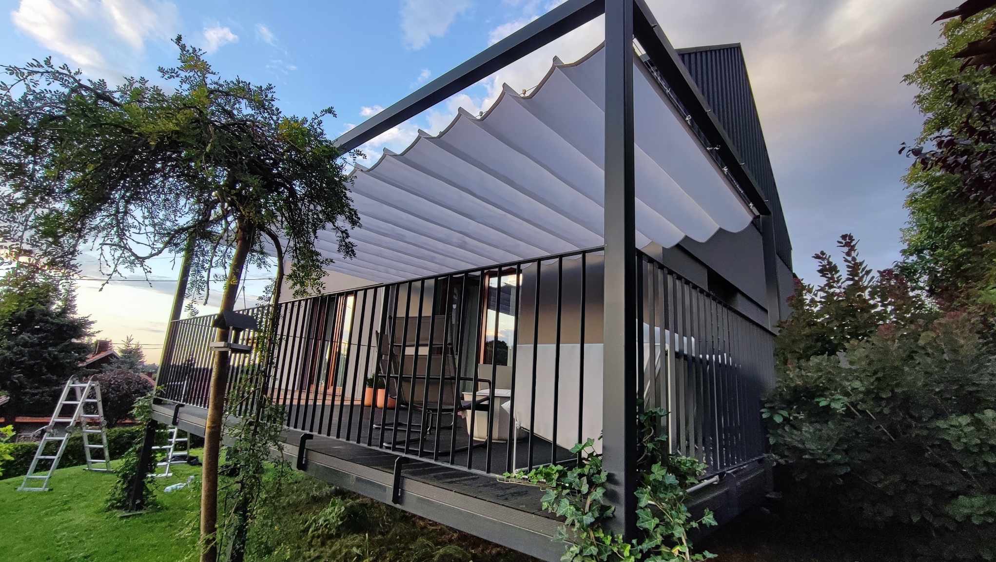 Nowoczesna pergola z falistym, białym zadaszeniem na balkonie domu w Strzelinie, widziana z dołu, z elementami zieleni w tle.