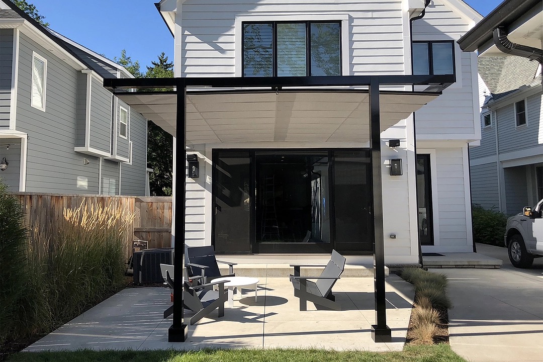 Nowoczesny taras z zadaszeniem z tkaniny na czarnej konstrukcji, z minimalistycznymi krzesłami i stolikiem, przylegający do białego domu z czarnymi oknami i drzwiami przesuwnymi.