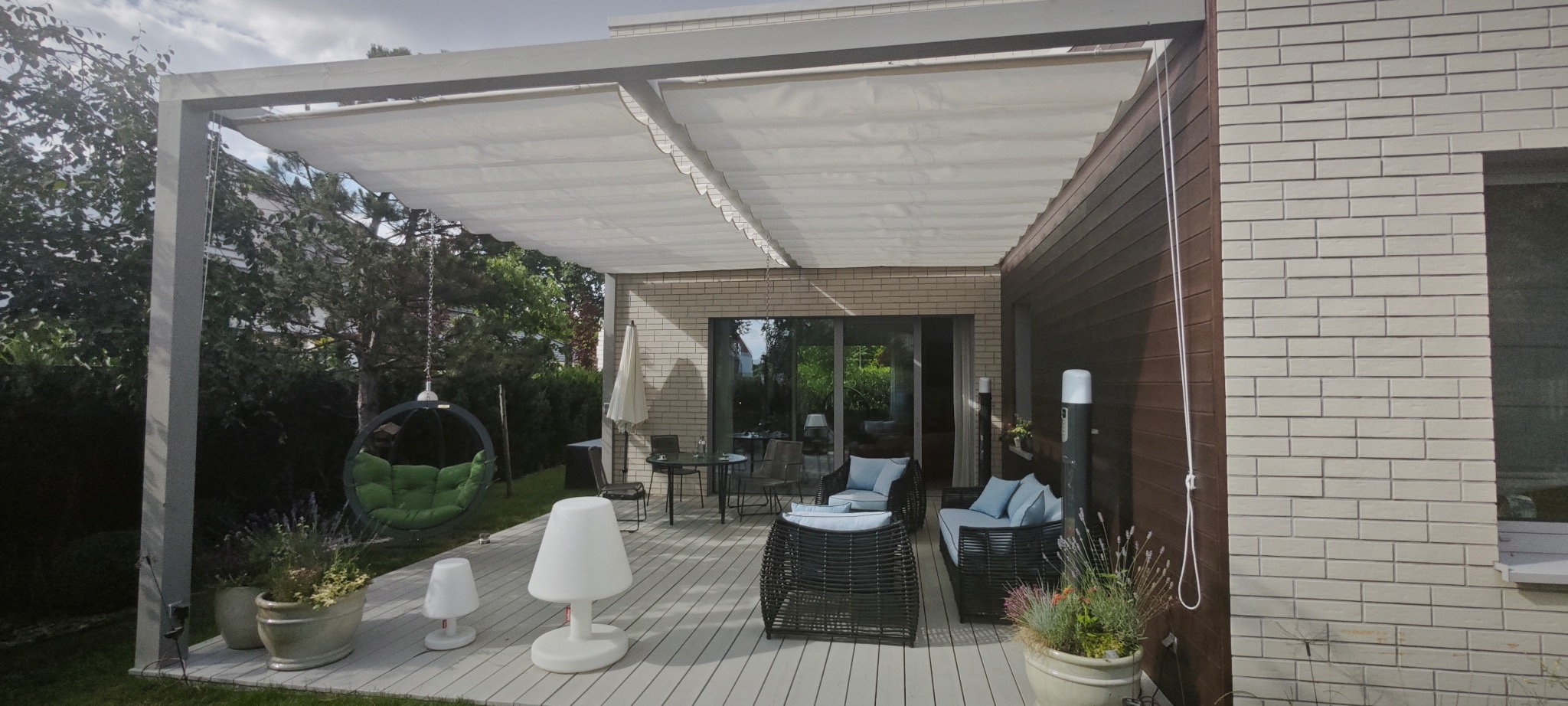 Nowoczesny taras z pergolą z jasną, zwijaną tkaniną, wiszącym fotelem i rattanowymi meblami wypoczynkowymi w pastelowych kolorach, oświetlony lampami podłogowymi.