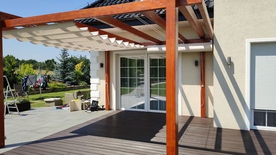 Drewniana pergola z beżową, rozsuwaną tkaniną zacieniającą taras przy domu z jasną elewacją i oknami z zamontowanymi zwiniętymi osłonami.