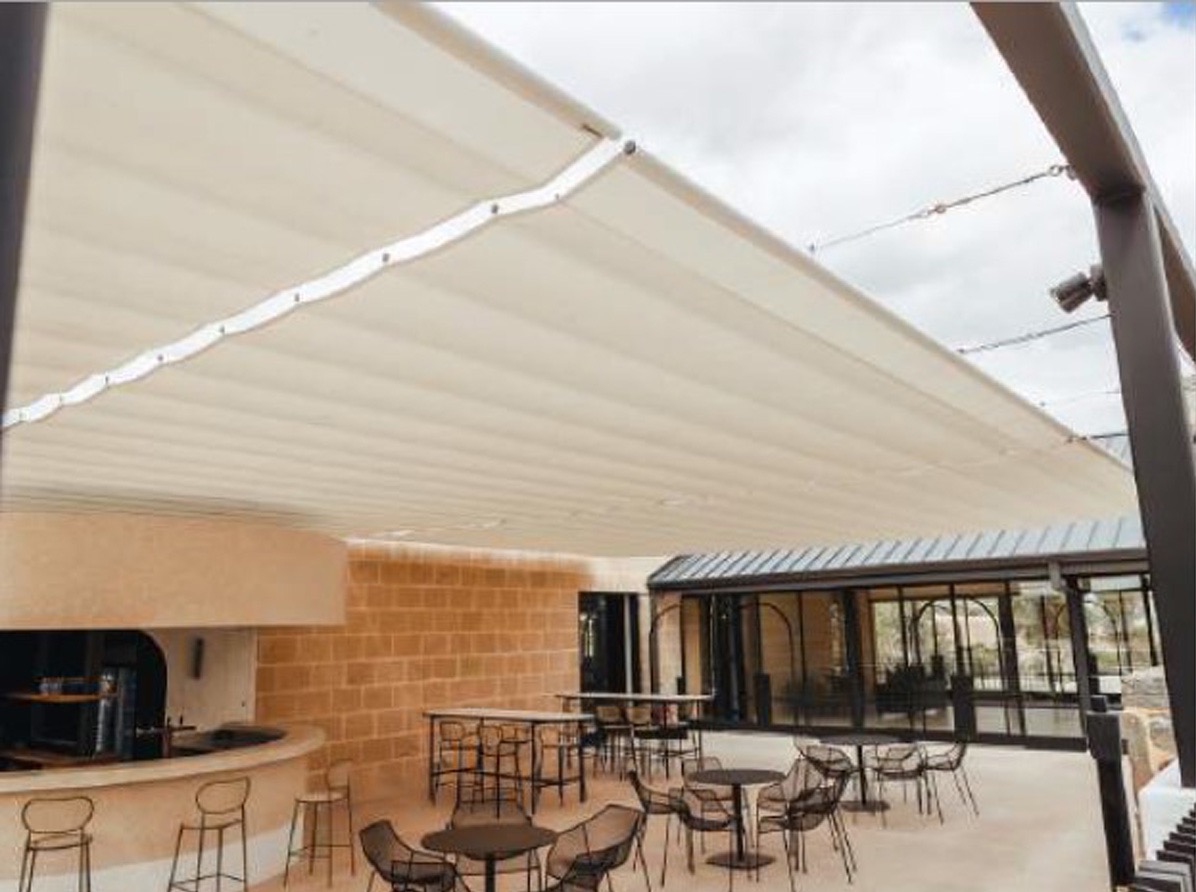 Beżowa, rozsuwana osłona przeciwsłoneczna nad tarasem restauracji z barowym stołem i krzesłami, widoczne metalowe słupy podtrzymujące konstrukcję.