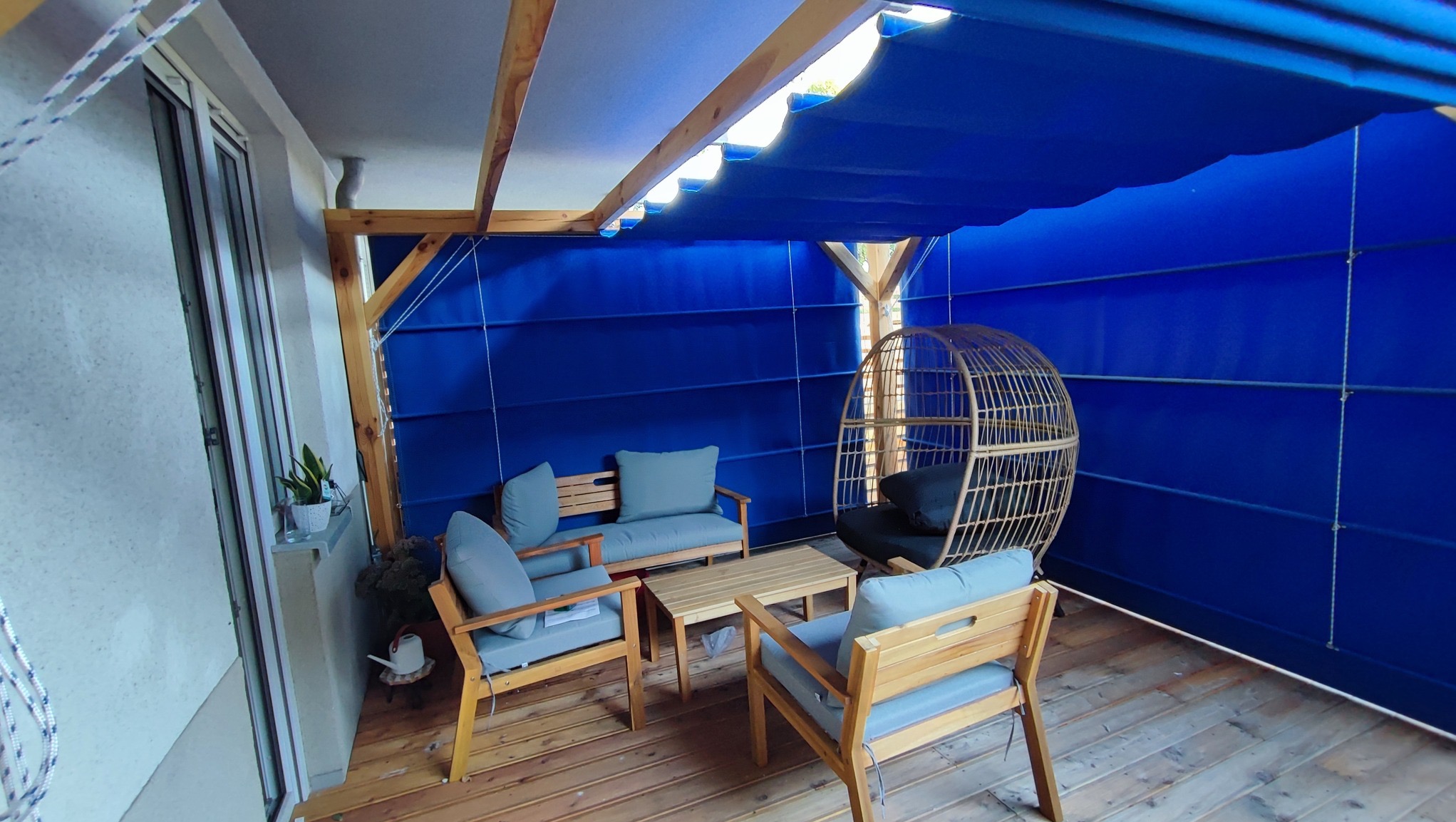 Zadaszony taras z drewnianą konstrukcją, niebieskim materiałem osłaniającym i meblami ogrodowymi z poduszkami w odcieniach szarości.