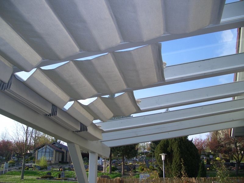 Widok z dołu na białą pergolę z częściowo rozłożoną, szarą tkaniną przeciwsłoneczną, przez którą widać błękitne niebo; w tle ogród z budynkiem.