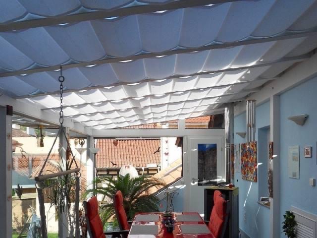 Zadaszenie tarasu z materiału falowanego, widok z wnętrza przeszklonej werandy, czerwone krzesła i stół w tle.