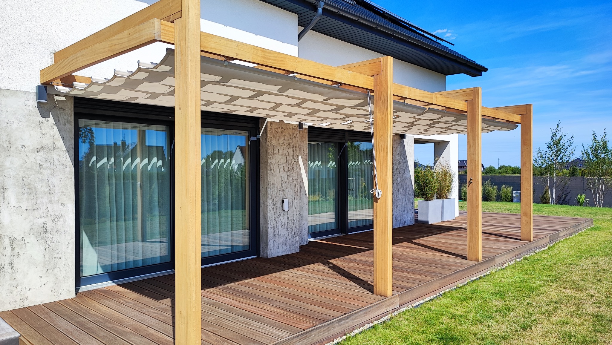 Nowoczesna pergola z drewnianą konstrukcją i rozsuwanym zadaszeniem materiałowym w geometryczny wzór, przylegająca do tarasu domu z dużymi oknami.
