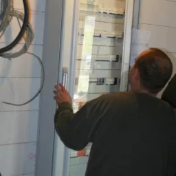 Otwarta szafa rozdzielcza z widocznymi zabezpieczeniami, instalowana na ścianie z bloków betonowych, oświetlona wiszącą lampą. Widoczne kable wchodzące do szafy od góry i od dołu, oraz fragment...