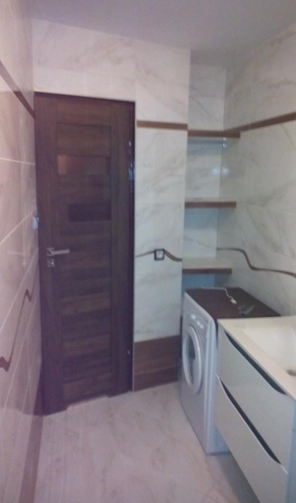 Wykończona łazienka w Kcyni z marmurowymi płytkami, drewnianymi akcentami i zabudową pralki obok umywalki.