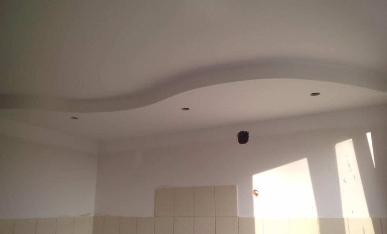 Wykończony sufit w kuchni z dekoracyjnym, falistym obniżeniem i otworami na oświetlenie punktowe, widoczne płytki na ścianie i ślady po instalacji elektrycznej.