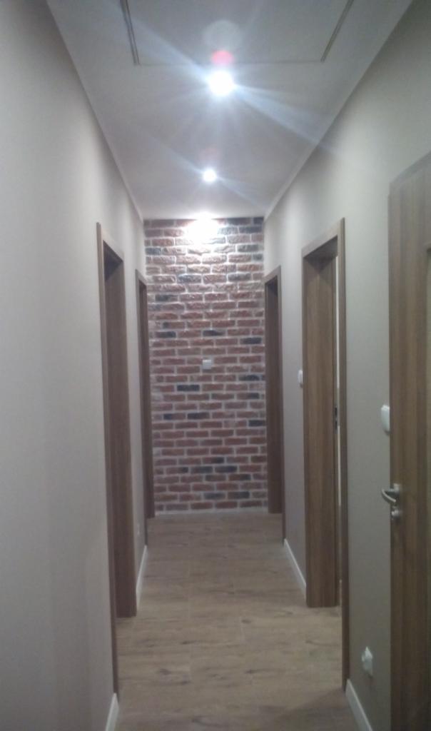 Wąski korytarz z ceglaną ścianą dekoracyjną, drewnopodobną podłogą i drzwiami z drewnianymi ościeżnicami, oświetlony punktowo.
