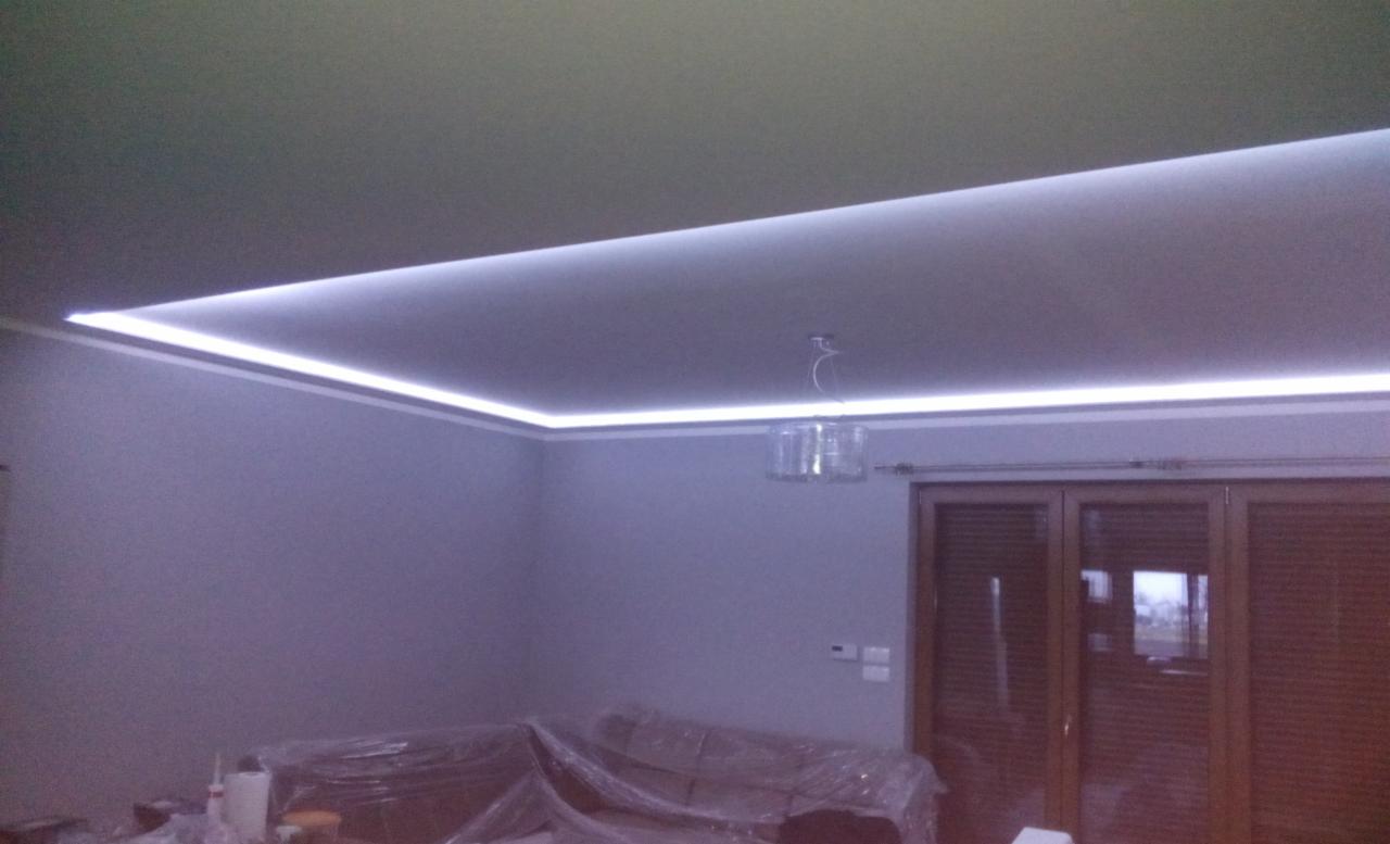 Nowoczesne oświetlenie LED zamontowane pod sufitem w rogu pokoju z widocznym żyrandolem i zasłoniętymi meblami folią ochronną.