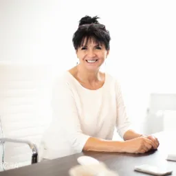 Uśmiechnięta kobieta z ciemnymi włosami siedzi za biurkiem w jasnym biurze, na biurku leży telefon i myszka komputerowa, w tle białe krzesło i monitor.