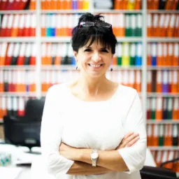 Uśmiechnięta kobieta w okularach, skrzyżowanymi rękami i zegarkiem na tle regału z segregatorami w biurze.