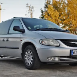 Srebrny Chrysler Grand Voyager z bagażnikiem dachowym, zaparkowany na betonowej nawierzchni z jesiennym drzewostanem w tle, widoczna tablica rejestracyjna WGR 14641.