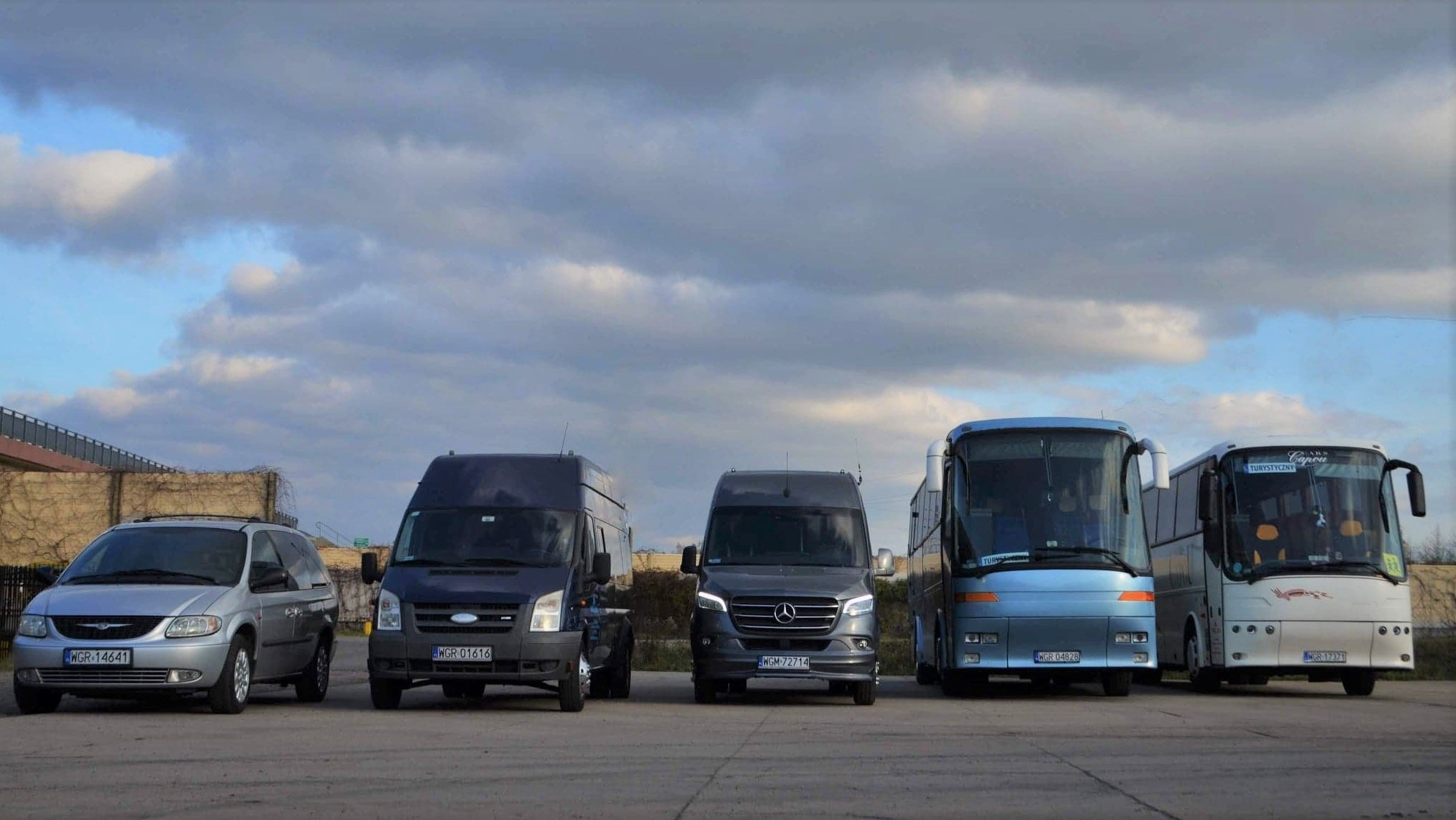 Rząd różnych pojazdów transportowych: srebrny minivan Chrysler, ciemny bus Ford Transit, szary Mercedes Sprinter oraz dwa autokary turystyczne na tle pochmurnego nieba.