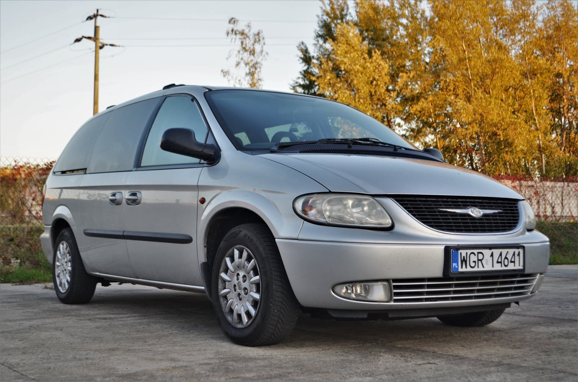 Srebrny Chrysler Grand Voyager z bagażnikiem dachowym, zaparkowany na betonowej nawierzchni z jesiennym drzewostanem w tle, widoczna tablica rejestracyjna WGR 14641.