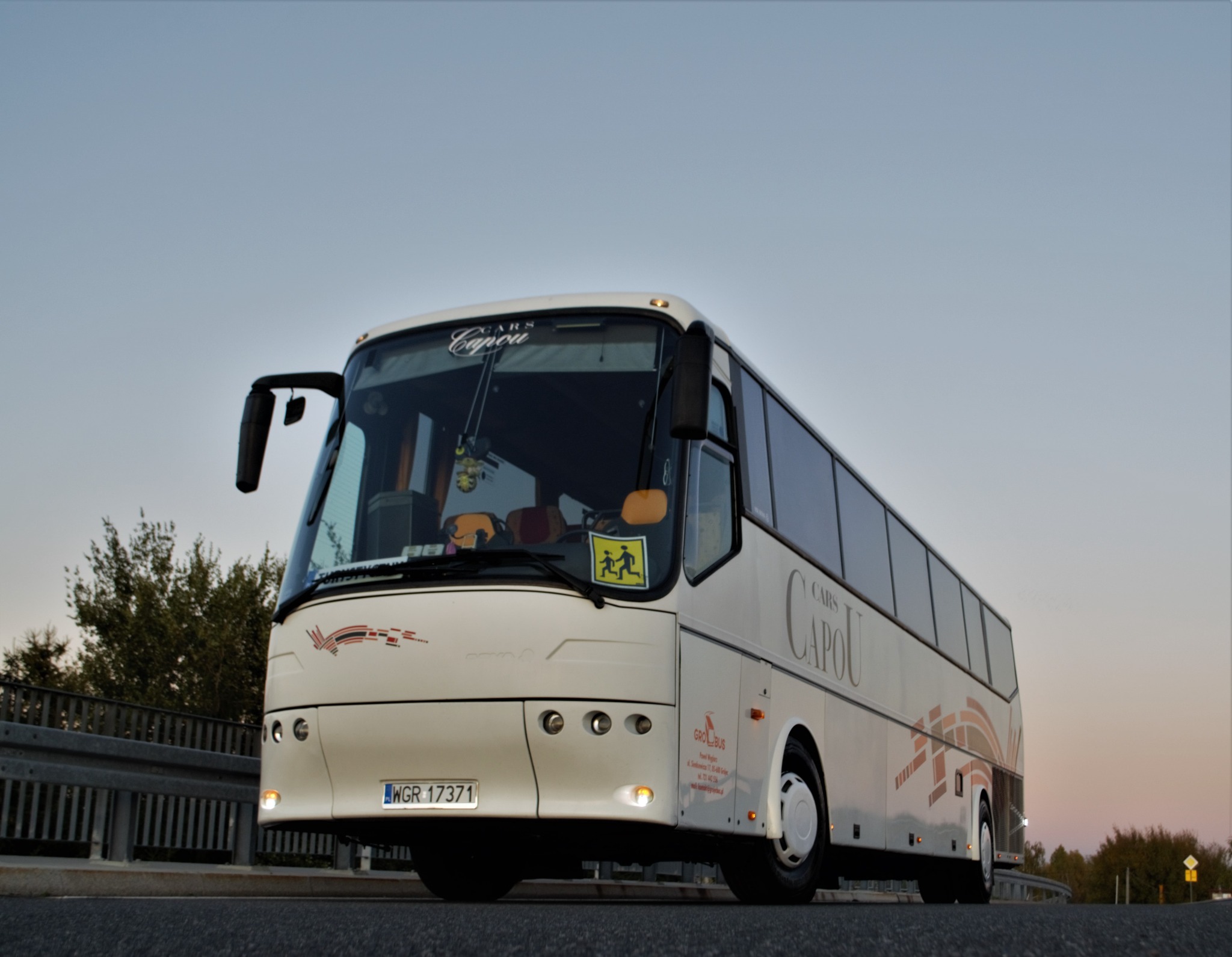 Biały autokar turystyczny marki BOVA VDL z napisem 'Cars Capou' na tle nieba o zmierzchu, widoczny znak ostrzegawczy 'dzieci'