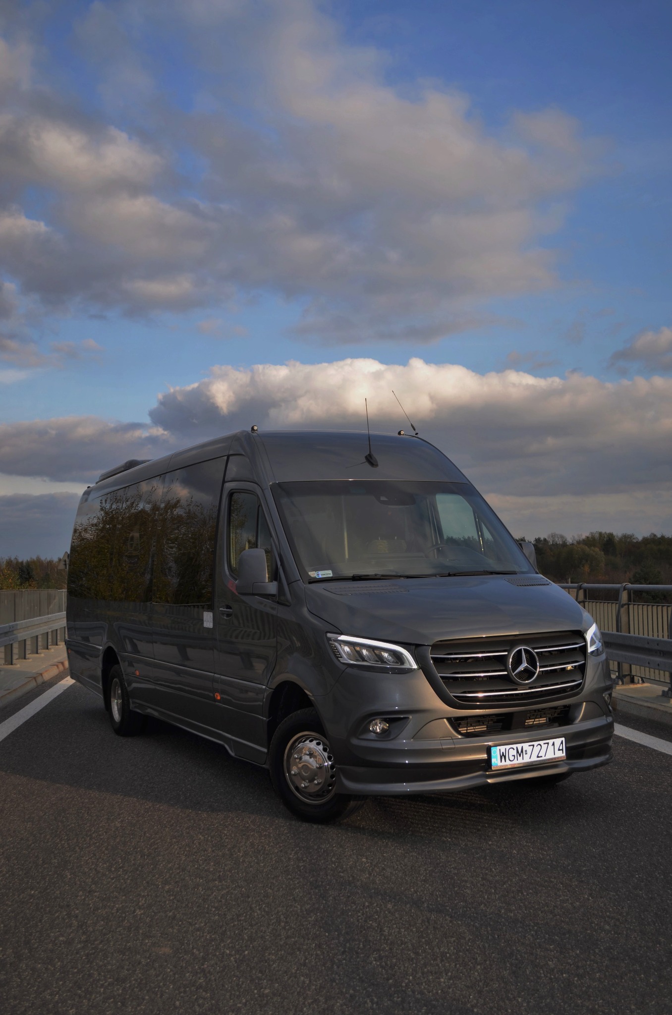 Szary Mercedes Sprinter VIP z antenami na dachu, zaparkowany na moście z widokiem na pochmurne niebo, odblaski na szybach.