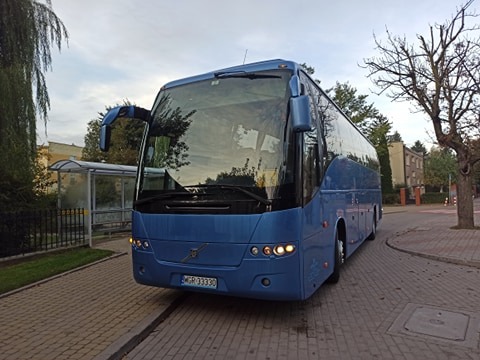 Niebieski autokar Volvo B12B z otwartymi drzwiami w przedniej części, zaparkowany na brukowanej drodze w pobliżu przystanku autobusowego i drzew.