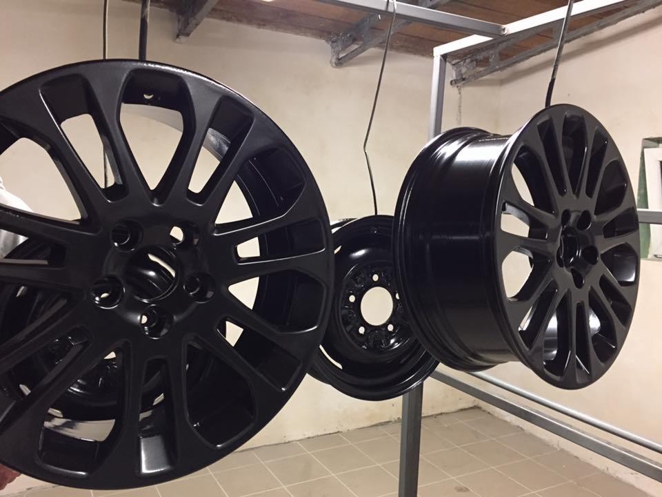 Trzy czarne felgi samochodowe zawieszone na metalowym stelażu w pomieszczeniu przypominającym lakiernię.