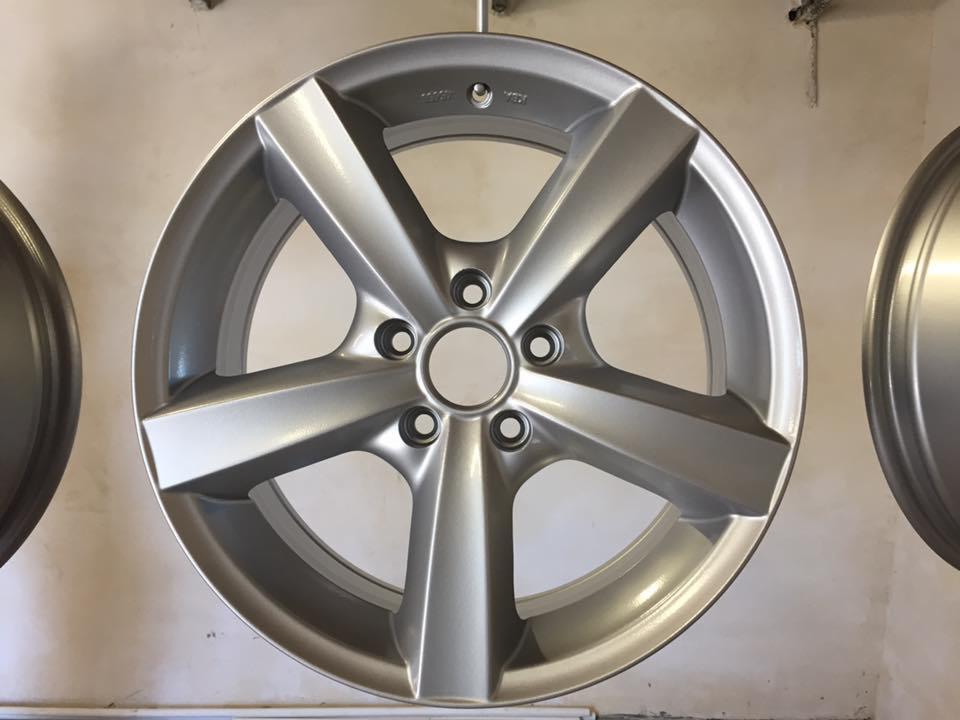 Świeżo pomalowana, srebrna felga aluminiowa o pięcioramiennej konstrukcji, zawieszona obok drugiej felgi, widoczna na tle jasnej ściany.