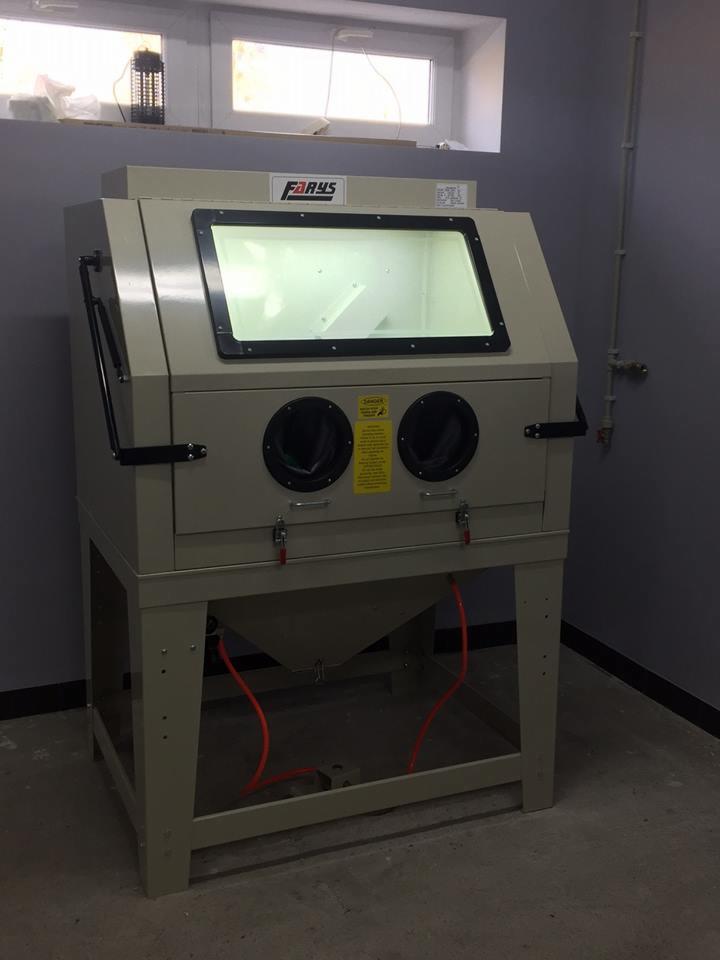 Stacjonarna piaskarka kabinowa z rękawicami i oświetleniem wewnętrznym, widoczny napis 'Farys' na obudowie, ustawiona w pomieszczeniu z oknem i instalacją na ścianie.