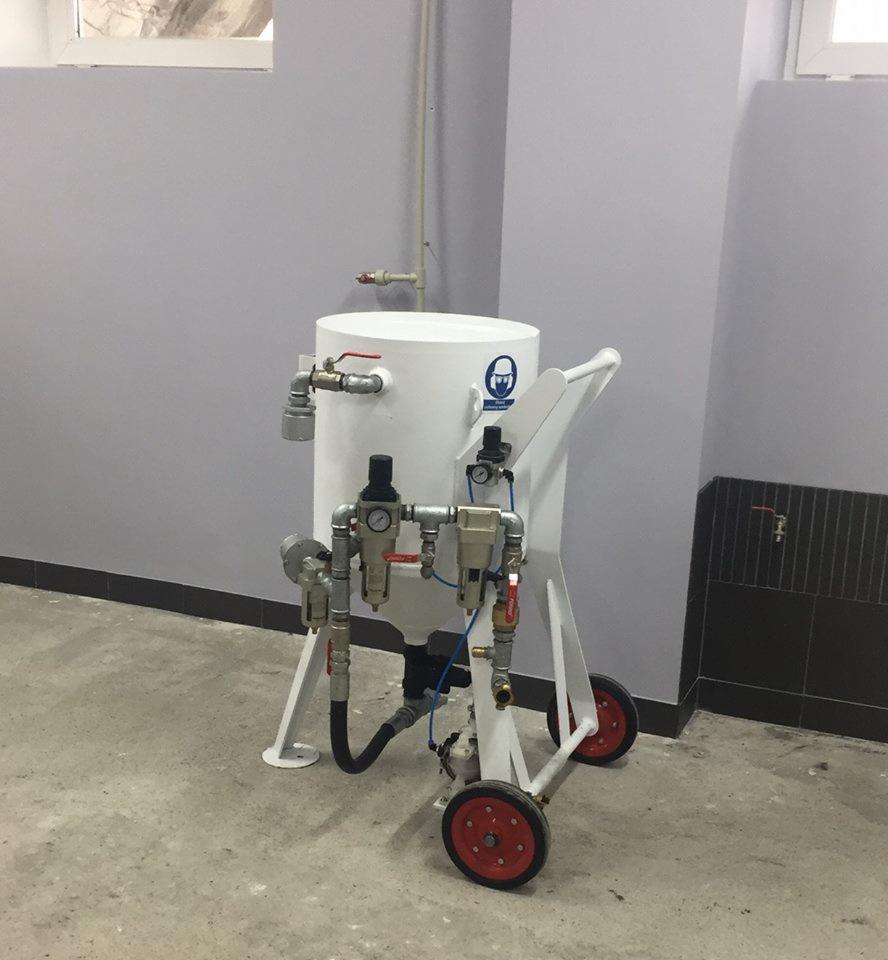 Mobilna piaskarka przemysłowa w białej obudowie z czerwonymi kołami, widoczna w pomieszczeniu o szarych ścianach i betonowej podłodze, wyposażona w liczne przewody i manometry.