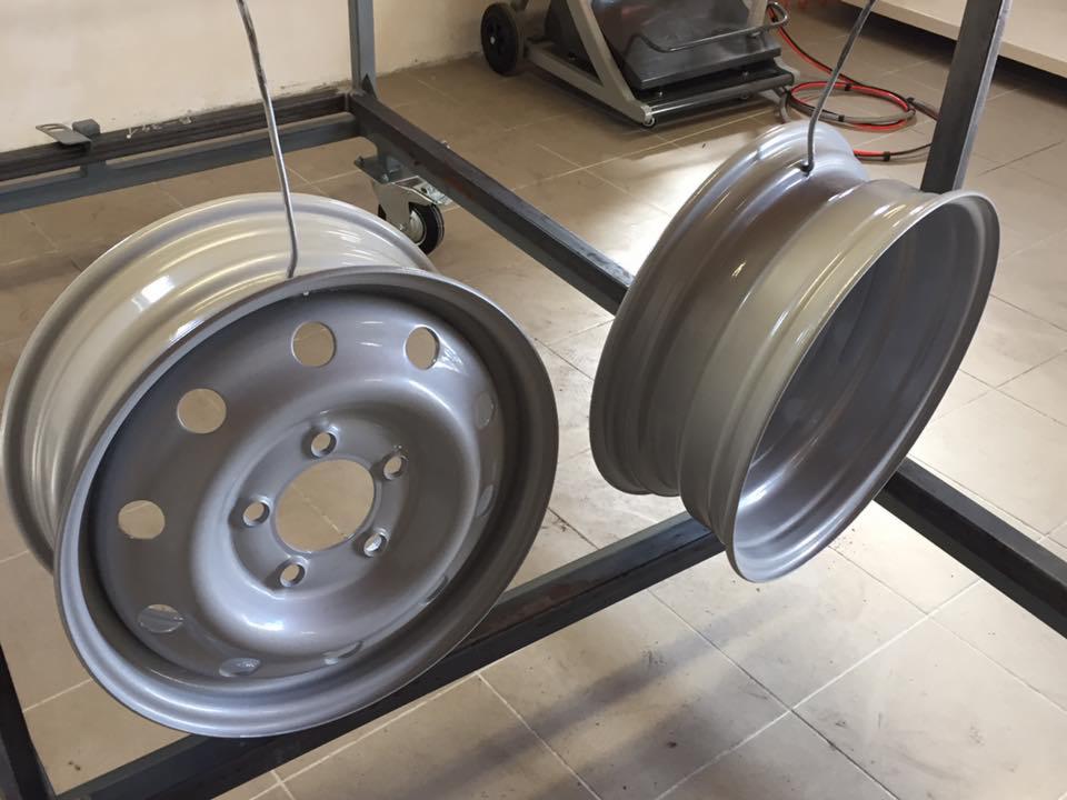 Dwie świeżo pomalowane na szaro felgi samochodowe zawieszone na metalowym stojaku, widoczny wózek z urządzeniem do malowania w tle.
