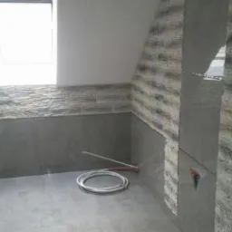 Łazienka w trakcie remontu, widoczne częściowo wykończone ściany z szarej, strukturalnej okładziny imitującej kamień, gładkie szare płytki na dolnej części ścian i podłodze, okno dachowe w skosie...